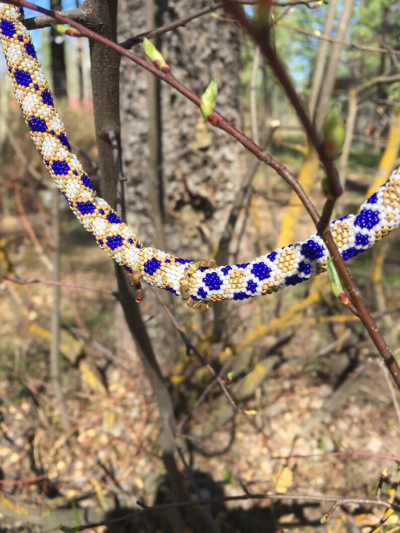 Puede incluir: Primer plano de un collar de cuentas azules, blancas y doradas. El collar est&aacute; envuelto alrededor de una rama con hojas verdes.