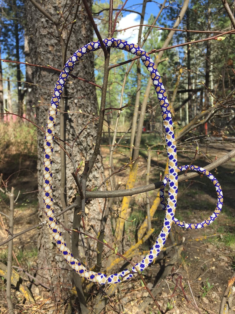 Puede incluir: Un conjunto de collar y pulsera de cuentas azules, blancas y doradas. El collar est&aacute; drapeado sobre una rama y la pulsera est&aacute; en el suelo.