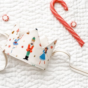 May include: A fabric crown with a festive Nutcracker print, featuring a ballerina, nutcracker, and other holiday elements. A red and white candy cane and peppermint candies are also present, along with a gold ribbon.