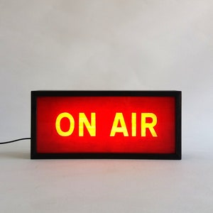 May include: A black framed sign with a red background and yellow text that reads "ON AIR".