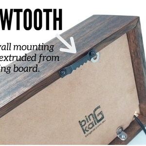 May include: A close-up of the back of a wooden frame with a sawtooth hanger for wall mounting. The hanger is made of metal and has a serrated edge. The frame is made of dark wood and has a brown backing board. The text "bing kai" is printed on the backing board.