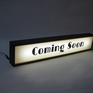 May include: A black framed sign with a white background and the text "Coming Soon" in black lettering. The sign is illuminated from behind.