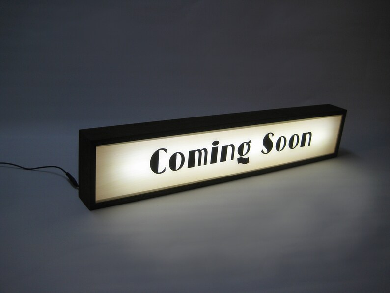 May include: A black framed sign with a white background and the text "Coming Soon" in black lettering. The sign is illuminated from behind.