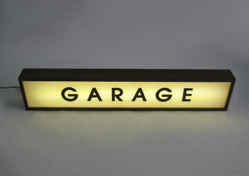 Pode incluir: Uma placa de madeira marrom com uma luz branca atr&aacute;s. A placa diz "GARAGE" em letras pretas.