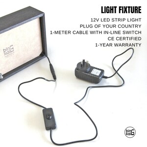 Pode incluir: Uma moldura de madeira preta com um adaptador de energia preto, um interruptor em linha preto e um cabo preto. A moldura tem um r&oacute;tulo branco com o texto "bing kali" nele. O texto "LIGHT FIXTURE" est&aacute; escrito em preto acima do texto "12V LED STRIP LIGHT". O texto "PLUG OF YOUR COUNTRY" est&aacute; escrito abaixo do texto "12V LED STRIP LIGHT". O texto "1-METER CABLE WITH IN-LINE SWITCH" est&aacute; escrito abaixo do texto "PLUG OF YOUR COUNTRY". O texto "CE CERTIFIED" est&aacute; escrito abaixo do texto "1-METER CABLE WITH IN-LINE SWITCH". O texto "1-YEAR WARRANTY" est&aacute; escrito abaixo do texto "CE CERTIFIED".