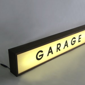 Pode incluir: Uma placa de madeira com um painel iluminado que diz "GARAGE" em letras pretas. A placa &eacute; iluminada por tr&aacute;s, criando um brilho quente.