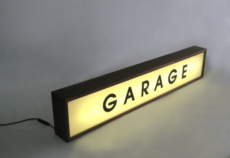 Pode incluir: Uma placa de madeira com um painel iluminado que diz "GARAGE" em letras pretas. A placa &eacute; iluminada por tr&aacute;s, criando um brilho quente.