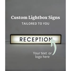 May include: A white lightbox sign with black lettering that reads "RECEPTION". The sign is illuminated and has a black frame. The text "Your text or logo here" is written below the sign.