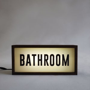 May include: A rectangular wooden-framed light box displaying the word "BATHROOM" in bold, black capital letters. The light box emits a warm, soft glow. A power cable extends from the left side. The logo "bin kai G" is in the bottom right corner.