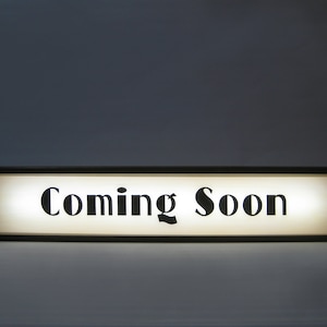 May include: A black framed sign with a white background and the text "Coming Soon" in black lettering.