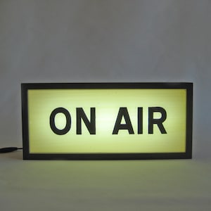 May include: A rectangular, illuminated sign with the words "ON AIR" in bold, black letters. The sign has a black frame and a soft, yellow glow emanating from within. A black cord extends from the side.