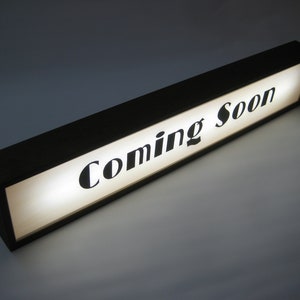 May include: A black wooden sign with a white background and the text "Coming Soon" in black lettering.