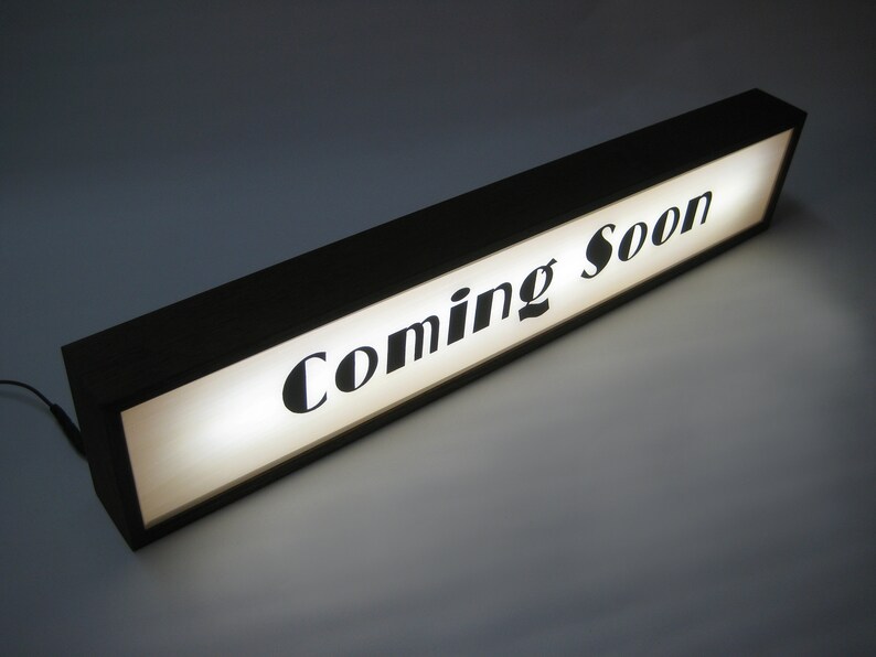 May include: A black wooden sign with a white background and the text "Coming Soon" in black lettering.