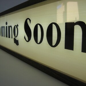 May include: A wooden framed sign with a white background and black lettering that reads "Coming Soon".
