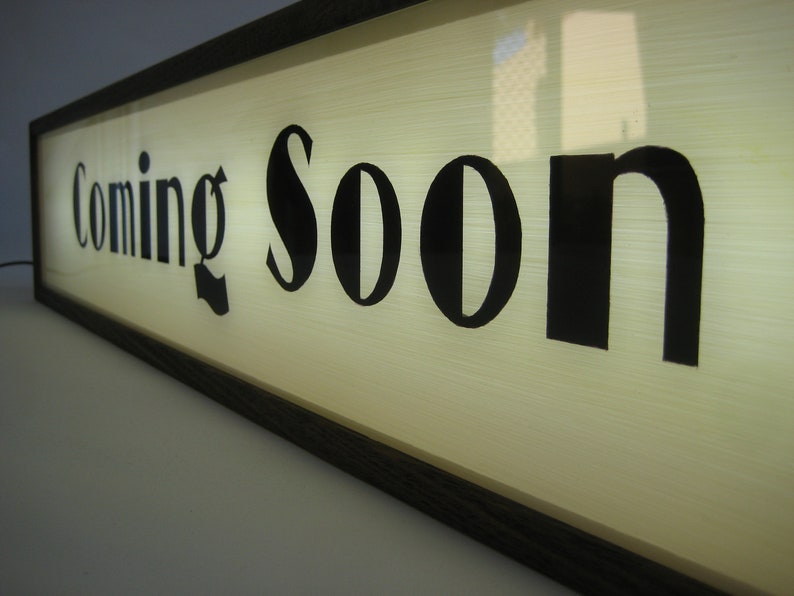 May include: A wooden framed sign with a white background and black lettering that reads "Coming Soon".