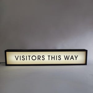 May include: A rectangular black sign with the words "VISITORS THIS WAY" in bold, white letters. The sign is illuminated from within, creating a bright, inviting display. The sign is designed to guide visitors.