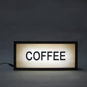 May include: A black framed light-up sign with the word "COFFEE" in white letters on a beige background.