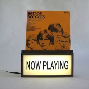 May include: A wooden record player stand with a light-up sign that says "NOW PLAYING". A vinyl record by the Bee Gees is displayed on the stand. The record cover is orange with black text and features a photo of the band members.
