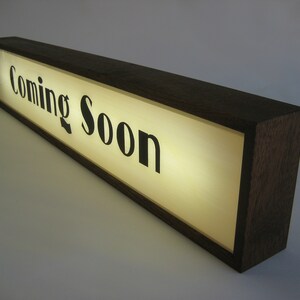 May include: A wooden sign with a lit-up panel that reads "Coming Soon". The sign is made of dark wood and has a rectangular shape.