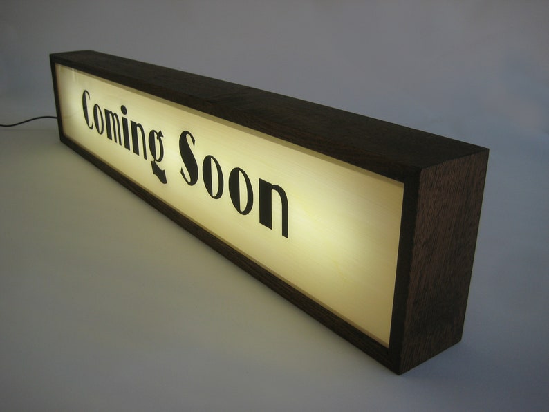 May include: A wooden sign with a lit-up panel that reads "Coming Soon". The sign is made of dark wood and has a rectangular shape.