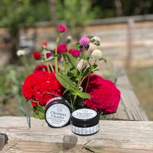 May include: Two black jars of activated charcoal toothpaste labeled "chomper cleaner" with a peppermint flavor. The jars are sitting on a wooden surface with red and pink flowers in the background.