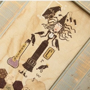 May include: A framed cross-stitch design featuring a witch holding a potion bottle. The witch is wearing a black dress and a pointed hat. The background is a brown and white patchwork design. The text "Madame Chan Tilly" is visible in the bottom right corner.