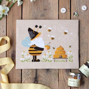 Puede incluir: Un bordado de punto de cruz con un gnomo con temática de abejas, una colmena y abejas. La obra de arte está sobre una superficie de madera con tarros de miel, narcisos y una cinta de cuadros amarillos. El texto "Bee Happy" está bordado encima de las abejas.