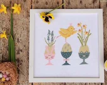 May include: A white framed cross-stitch design featuring a yellow chick standing on an egg with a bunny face, a blue egg with a bow tie, and a cat egg. The eggs are in a vase with flowers.