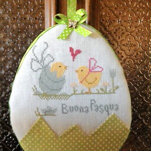 May include: A white egg-shaped fabric ornament with green polka dot trim. The ornament features an embroidered design of two birds and the text "Buona Pasqua".