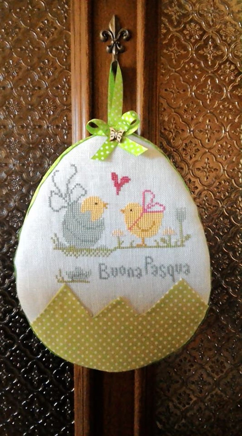 May include: A white egg-shaped fabric ornament with green polka dot trim. The ornament features an embroidered design of two birds and the text "Buona Pasqua".