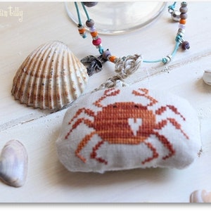 May include: A white fabric crab ornament with orange and brown embroidery. The ornament is hanging from a blue and white beaded necklace. There are several seashells on a white wooden surface.