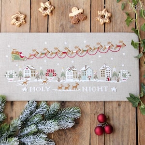 Op de afbeelding: Een kruissteekontwerp op beige stof toont een kerstscène met de slee van de kerstman getrokken door rendieren boven een dorp. De woorden "HOLY NIGHT" zijn eronder geborduurd. Kerstversieringen omringen het beeld.
