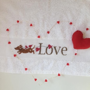 Puede incluir: Toalla blanca con un dachshund bordado en marrón corriendo con corazones rojos. La palabra "LOVE" está bordada en marrón debajo del dachshund. Una cadena de luces de corazón rojo está dispuesta en forma de corazón alrededor del bordado. Un corazón de fieltro rojo está en la esquina superior derecha.
