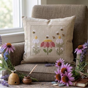 Può includere: Un cuscino quadrato color beige con un ricamo a punto croce raffigurante tre fiori e diverse api. Il fiore centrale è rosa con il centro giallo, affiancato da due fiori bianchi. La parola "sweet" è ricamata in bianco sopra i fiori.