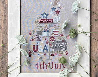 May include: A framed cross-stitch embroidery featuring a tiered stand with patriotic motifs, including an eagle, American flags, and the text Freedom, USA, Liberty, and 4th of July, displayed in a light-colored wooden frame with white floral accents.