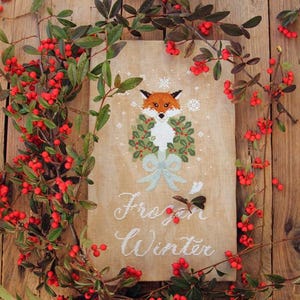 Puede incluir: Un diseño de punto de cruz con un zorro con una corona de acebo, con las palabras "Frozen Winter" debajo. El diseño está rodeado por una guirnalda de bayas rojas y hojas verdes, sobre un fondo de madera.