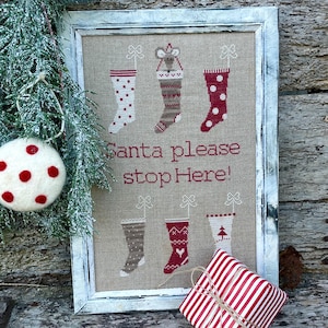 May include: A framed cross-stitch design featuring six stockings with different patterns and the text "Santa please stop here!"  A red and white striped gift box is in the foreground.