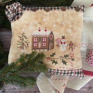 May include: A beige cushion with a cross-stitch design of a gingerbread house, a snowman, and a holly bush. The cushion has a plaid border and is decorated with buttons.