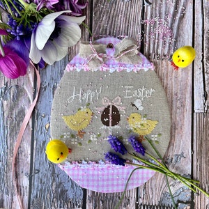 Może przedstawiać: Haftowany krzyżykiem wzór przedstawiający różowo-białe jajko w kratkę z napisem "Happy Easter" i trzema żółtymi kurczakami. Jajko otoczone jest fioletowymi kwiatami i różową wstążką.