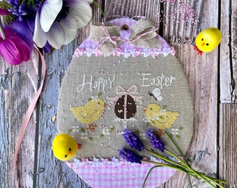 May include: A cross-stitch embroidery of a pink and white gingham egg with the text "Happy Easter" and three yellow chicks. The egg is surrounded by purple flowers and a pink ribbon.