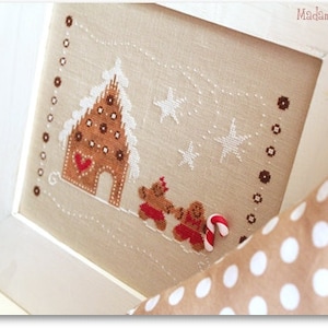 May include: A cross-stitch embroidery design featuring a gingerbread house with a red heart, two gingerbread people, and white stars on a beige background. The design is framed in a white frame.