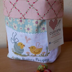 May include: A white fabric Easter basket with a pink, blue, and white floral pattern. The basket is filled with chocolate eggs wrapped in foil. The basket has a cross-stitched design of two chicks and the words "Buona Pasqua". The basket has a tag attached that says "Cuorarte by Sonia Vecchi, ricamo e cucito passione".
