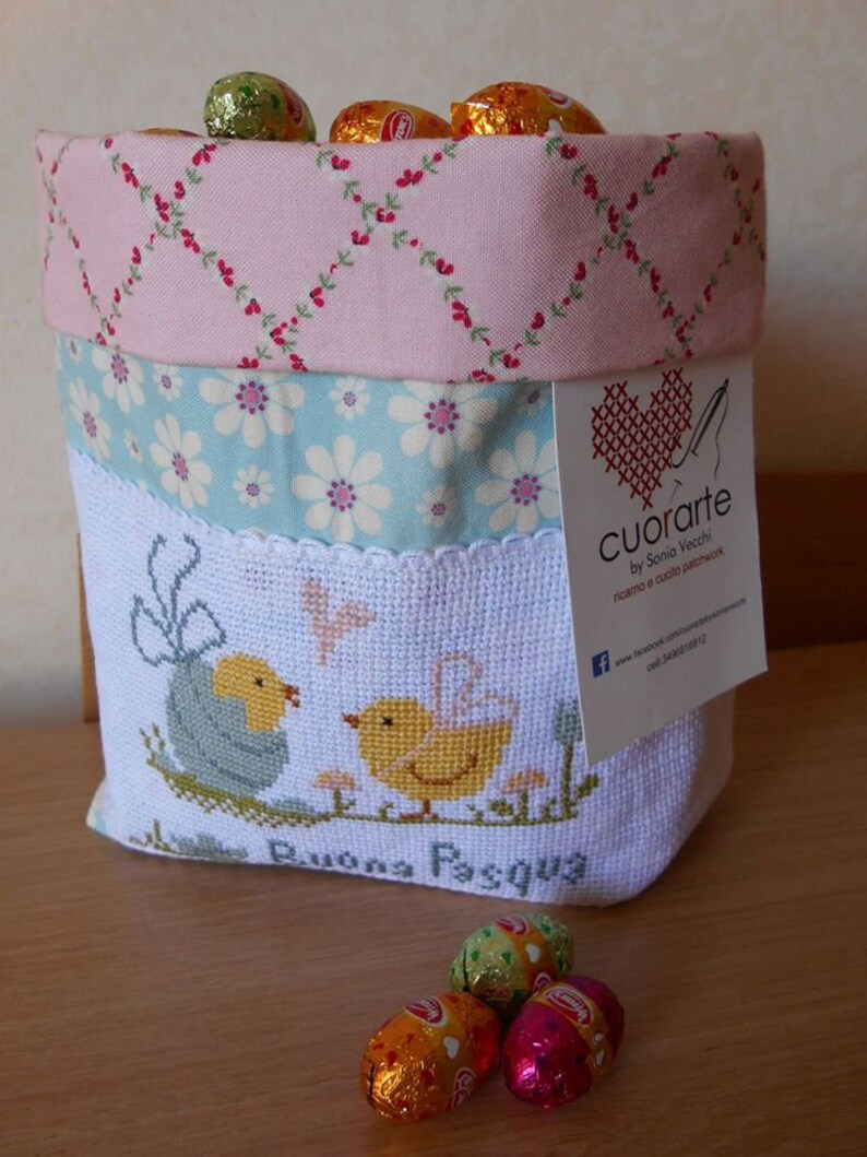 May include: A white fabric Easter basket with a pink, blue, and white floral pattern. The basket is filled with chocolate eggs wrapped in foil. The basket has a cross-stitched design of two chicks and the words "Buona Pasqua". The basket has a tag attached that says "Cuorarte by Sonia Vecchi, ricamo e cucito passione".