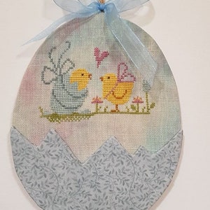 May include: A blue and white fabric Easter egg ornament with a cross-stitch design of two yellow chicks with wings and a pink butterfly. The egg is decorated with a blue ribbon and a brown twine hanger.