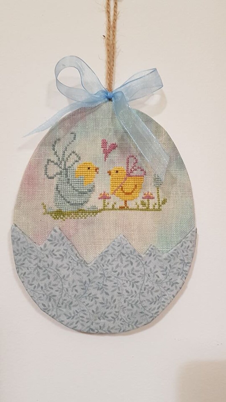 May include: A blue and white fabric Easter egg ornament with a cross-stitch design of two yellow chicks with wings and a pink butterfly. The egg is decorated with a blue ribbon and a brown twine hanger.