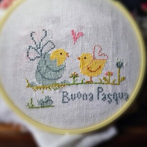 May include: A cross-stitch embroidery hoop with a white background featuring two chicks and a butterfly. The text "Buona Pasqua" is stitched in the bottom center of the hoop.