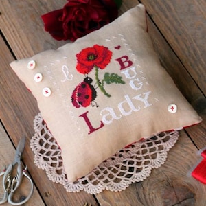 Puede incluir: Un cojín cuadrado beige con borde rojo, bordado con una amapola roja, una mariquita y las palabras "I Bug Lady". El cojín está adornado con botones blancos y descansa sobre un tapete de encaje. Una rosa roja y un pequeño bastidor de bordado están cerca.