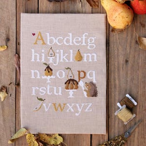 Puede incluir: Un muestrario de punto de cruz con el alfabeto, con motivos de pera y erizo. Las letras son blancas y doradas sobre tela beige. Peras y hojas esparcidas sobre una superficie de madera. Tijeras doradas.