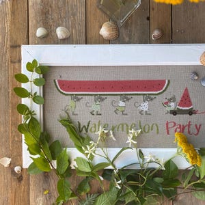Op de afbeelding: Een ingelijst kruissteek borduurwerk op lichtbruine stof met vier muizen die een groot watermeloenpart boven hun hoofd dragen, een vijfde muis trekt een wagentje met een watermeloenpart, en de tekst Watermelon Party in groen en rood.
