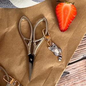 Puede incluir: Un par de tijeras pequeñas plateadas con un llavero de gato dorado. El llavero de gato tiene un gato gris y blanco con ojos negros y una lengua rosa. Las tijeras y el llavero están sobre una superficie de papel marrón.